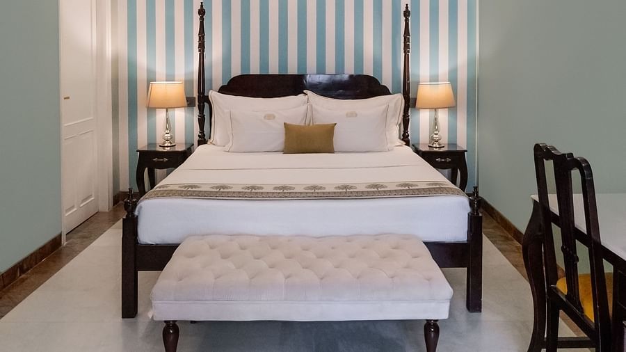 A narrow bedroom with a four-poster bed, vertical striped wallpaper, pale green walls, and a white floor, with a bench and chairs on either side | Dileep  Kothi, Jaipur